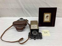 Three items displayed: framed lady portrait on porcelain, Pickwick Candid type camera with instruction manual, and Voigtlander camera in leather case.