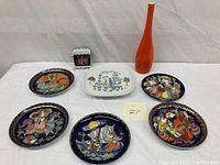 Group photo showing six decorative collector plates, one orange slender vase, and one small white floral square vase arranged on a white surface.