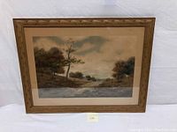 Framed lithograph artwork showing a landscape scene with trees and a river or path under a cloudy sky.