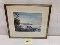 Frontal view of the framed watercolor painting showing seascape with rocks and trees. Displayed with lot number.