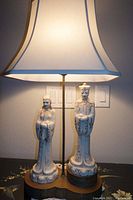 Wide shot of lamp showing porcelain figurines of man and woman, brass base, and silk lamp shade illuminated.
