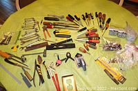Photo showing assortment of hand tools including wrenches, screwdrivers, pliers, ratchets, adjustable wrenches, saw, measuring tape, and bags of bolts and screws on a green tablecloth.