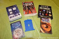 Full lot view showing five Doctor Who books arranged in a circle on a green fabric and the blue TARDIS wall light switch cover in the center.