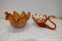 Two amber/orange glass bowls placed side by side on fabric. One is a ruffled handkerchief-style bowl, and the other is swan-shaped with shells inside.