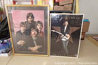 Two framed posters leaning on the floor, one large Beatles poster with four men, the other Paul McCartney tour poster with him holding a guitar.