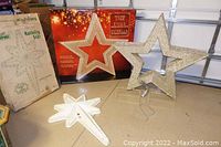 Two large gold tinsel star decorations approximately 39-40" tall with their original boxes and metal stands placed on a concrete floor. One star is upright, the other star's section with plastic picks and base is shown.