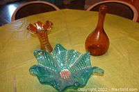 Photo showing all three items on a green textured tablecloth. Includes an amber tall vase with narrow neck and round base, an amber carnival glass vase with ruffled top and vertical ribs, and a blue wavy-edged glass bowl with textured bottom.