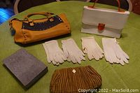Overview of three purses (yellow/gray leather handbag, white vinyl box handbag with amber lucite handle, greenish-brown velvet clutch) along with four pair white gloves and original box on green surface.
