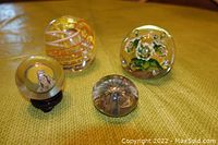 Four vintage glass paperweights arranged on yellow-green fabric surface, showing all items clearly with varying designs including floral, spiral, and painted figure.