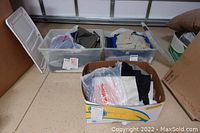 Plastic bins and a cardboard box filled with folded men's slacks, jeans, khakis, and shorts in a variety of colors, many new with tags.