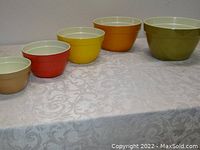 Five nested melamine mixing bowls in olive, orange, yellow, red, and tan colors arranged in a row on a patterned cloth surface showing their size difference.