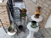 Wide view of assorted metal and pedestal-style plant stands with decorative watering cans and the black metal wine rack on a porch.