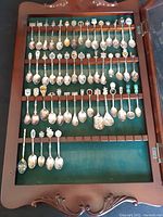 Front view of spoon display case with spoons inside showing various collectible designs and green felt background.