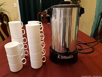 Photo showing the West Bend coffee urn with electric cord next to 18 stacked white coffee cups on a table.