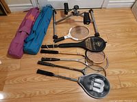 Lot of two Lotus yoga mats in purple and teal with bags, pedal exerciser, and six rackets laid out on wooden floor.