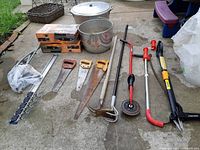 Wide angle including all items in the lot spread out: saws, garden tools, pots, boxed lanterns, and hand pump