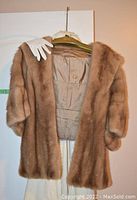 The pale mink fur stole hanging on a hanger with a pair of white gloves draped over it. The lining with a gold emblem inside is partially visible.