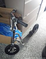 Side angle showing metal silver frame, black handle grips, black deck, two large black tires, front one flat, hand brake on handlebars.