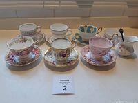Overview of six sets of bone china tea cups and saucers with sugar and creamer on countertop.