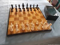 Full view of vintage wooden chessboard with all chess pieces set up in starting positions plus a black storage box on table.