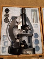 Top view of black vintage Tasco microscope neatly set in original foam fitted box with three additional eyepieces visible.