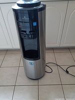 Full view of the cylindrical stainless steel Vitapur water dispenser standing on tile floor, plugged in with power cord visible.