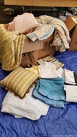 Overview of assorted bed linens, blankets, pillows, and dust ruffles arranged on and around a cardboard box on a blue tarp.