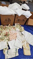 Boxes filled with white bed linens spilling out including sheets and pillowcases, some floral and striped fabric remnants visible in front
