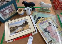 Overview of lot including both cast iron sheep doorstops, ceramic mugs and pitchers, acrylic place mats in box, decorative tray, and framed artworks.