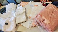 View of various folded linens including white embroidered pieces, peach colored linens, and packaged towels all laid on table.