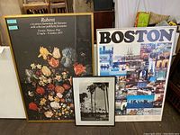 Three framed items leaning against wall: Rubens floral poster, Boston collage poster, black-and-white palm tree photo