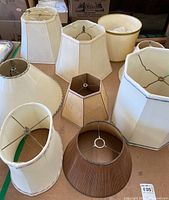 Nine assorted lamp shades arranged on a table showing different shapes and sizes, mostly cream colored fabric with metal frames.
