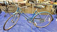 Blue mid-century women's Schwinn bicycle with step-through frame, black saddle, curved handlebars, showing rust and wear on rims, chain, and metal components, on blue tarp.