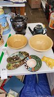 Photo of vintage pottery items on white table including two teapots, two yellowware bowls, ceramic pig, painted ashtray, and painted slabs.