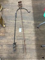 Two garden shepherd's hook posts laid flat on wooden floor, one black metal with clamp, one rusted with decorative scroll and plate top.