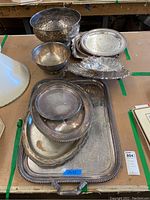 Overview of 11 silver-plated trays and bowls displayed on a table, showing various shapes, sizes, and decorative edges.