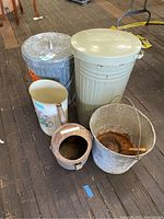 Image showing five vintage metal items: two trash cans, one cream-colored and one galvanized steel; milk bucket with faded red 'Security Food' label; rusty coal ladle with wooden handle; floral watering can.