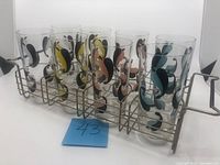 Front angled view of all 8 glasses in wire glass holder rack showing colorful patterns on clear glasses and rack details.