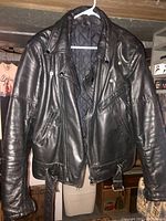 Front view of black leather motorcycle jacket hanging on a plastic hanger indoors, showing zipper pockets and belt detail at bottom.