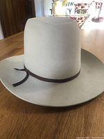 Front and side angled view of beige Western cowboy hat featuring a brown band tied in a knot, sitting on wooden surface.