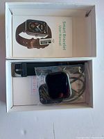 Smart bracelet in box with user manual visible and charging cable packaged separately in plastic.