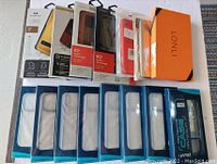 Photo showing approximately 15 brand new phone cases in original packaging, including blister packs with blue background and branded boxes such as 'LONLI'.
