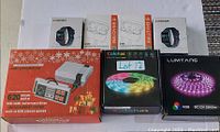 Photo of the lot containing two Lbstar E90 smart watches in boxes, a retro mini game console with a controller, and two RGB LED strip light sets in black boxes.