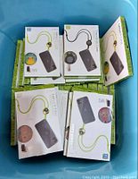 Photo shows approximately 40 boxed new crossbody and lanyard phone cases in an open container. Cases have lime green and black lanyards with small charms. Boxes display image of phone case with lanyard and a smartphone.