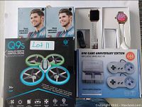 Photo shows two boxes of True Wireless Headsets, a boxed Q9S drone, a Mini Game Anniversary Edition console with two wired controllers, and a white smartwatch with rose gold case and perforated strap.