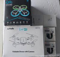 Photo showing Q9s drone box, Zuhafa foldable drone box, two L8Star smart watch boxes, and one wireless earbuds box on a gray surface.