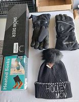 Photo shows the contents of the lot: a box of Snow Deer heated gloves, the gloves themselves laid out, a box of heated socks for foot warming, and a black knitted Hockey Mom touque with a fur pom-pom.