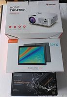 Photo showing three boxed electronics items stacked: Bomaker Home Theater Projector C9 (white projector), Vankyo MatrixPad S30 Android Tablet (gray tablet), and Bogasing M4 Wireless Speaker (black speaker box).