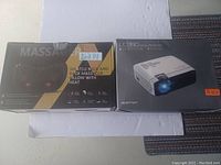 A photo showing the boxed Shiatsu neck and back massager pillow with heat and the Optoma LC330 digital projector box side by side.