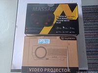 Photo of Vankyo video projector in cardboard box and Shiatsu neck and back massager pillow with heat retail box stacked on top.